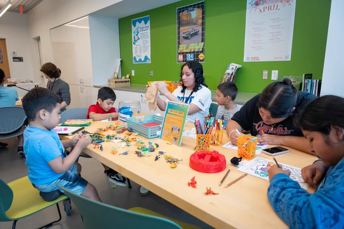 A group of children and a facilitator gather around a table for a craft activity in a bright room with green walls. Some kids color while others assemble small projects. Paper, markers, and craft supplies are scattered across the table, with posters visible on the surrounding walls.