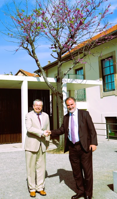 Two men in business suits shake hands outdoors in front of a modern building with white walls and large windows. A blooming tree with pink flowers stands behind them under a clear blue sky