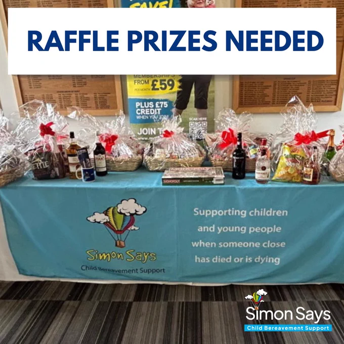 Photograph of a table with a blue Simon Says CBS table cloth covering it. On the table are a wide selection of raffle prizes. There are hampers which are wrapped and decorated with red bows. There are also bottles of wine, bottles of spirits and a board game. Text imposed over the photograph reads “Raffle prizes needed.”