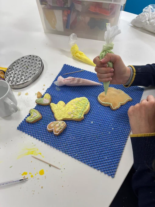 Biscuits in the shape of hearts and bunnies are on a blue mat being iced using a piping bag filed with pale green icing. 