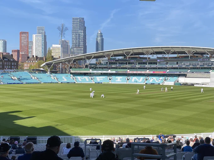 Cricketers in their whites playing at the Kia Oval with clear blue sky behind the grandstand