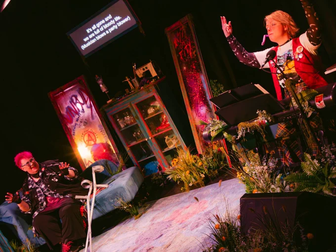 Production shot of Unruly. Shows two female performers either side of a stage. Both are dressed in punk outfits. The set includes mirrors with disability rights slogans written on them