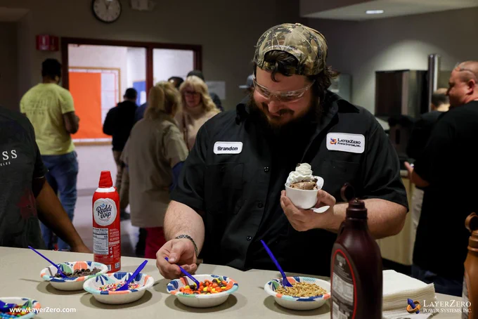 This image features a smiling man holding a white, plastic bowl in the shape of a baseball cap filled with chocolate ice cream, whipped cream, and chocolate chips. He is scooping candy onto his ice cream. There are four bowls of different toppings to choose from and a can of whipped cream and chocolate sauce. He is wearing a backwards LayerZero hat and a LayerZero button-up shirt that reads “Brandon” on the left and has the logo on the right. The bottom left corner has text that reads www.LayerZero.com. The bottom right corner has the LayerZero logo.