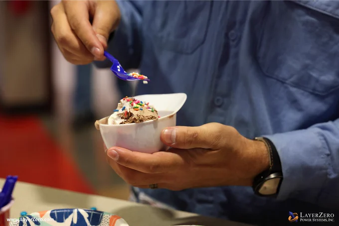 This image features a close up of a man holding a white, plastic bowl in the shape of a baseball cap with chocolate and vanilla ice cream in it. He is pouring multi-colored sprinkles on top of the ice cream. The bottom left corner has text that reads www.LayerZero.com. The bottom right corner has the LayerZero logo.