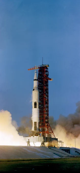 A white rocket lifts off from the launchpad. An orange gantry can be seen right next to the rocket. White vapor spreads outward on the left and right sides of the image. The vapor is lit up by flames from the rocket. Credit: NASA