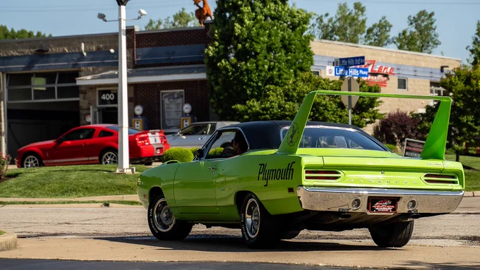 A bright green Plymouth muscle car with a large rear fin is parked in front of a commercial building. A red car is visible further back in the scene, with trees and a blue sky in the background. The setting suggests an automotive or classic car event.