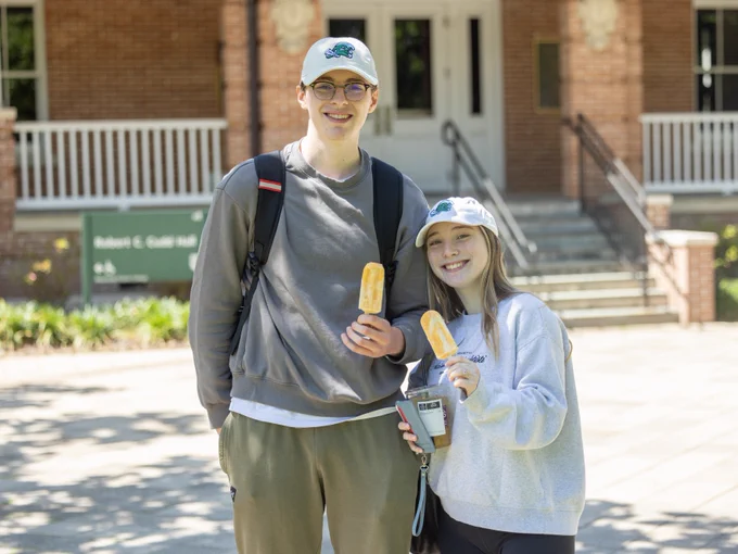 Two students holding ice cream bars in front of a brick building on Tulane University's campus, smiling at the camera.
