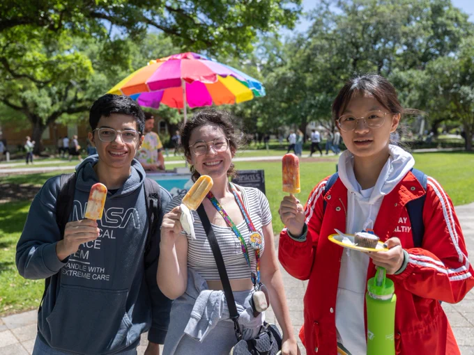 Three individuals holding popsicles under a colorful umbrella on a sunny day at an event on campus.