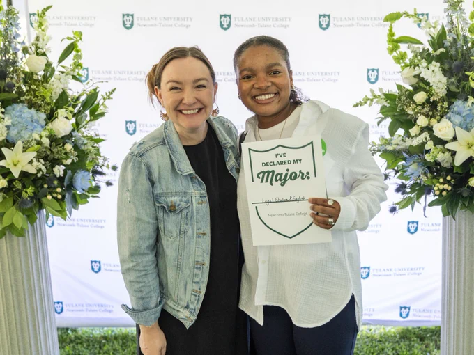 Two individuals smiling at a Tulane University event, one holding a sign that reads "I've Declared My Major!" with floral arrangements and a banner with the university logo in the background.
