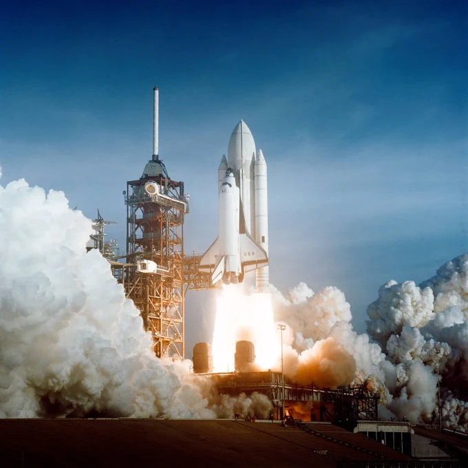 The space shuttle roars to life as it lifts off the launchpad for the first time. The white and black airplane-shaped orbiter is attached to a large white fuel tank and two solid rocket boosters. Bright flames are seen from the boosters and engines as the space shuttle leaves the launch pad and rises next to the launch tower
