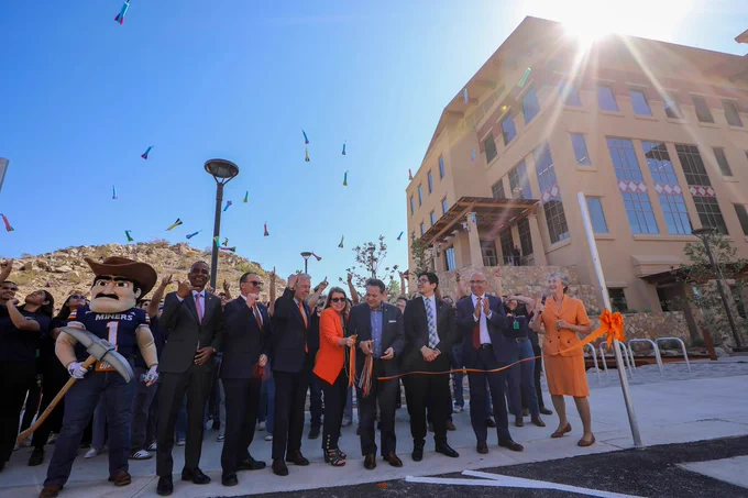 A group of people and the UTEP Paydirt Pete mascot cut an orange ribbon while standing in front of a building. Toy rockets fly in the air.