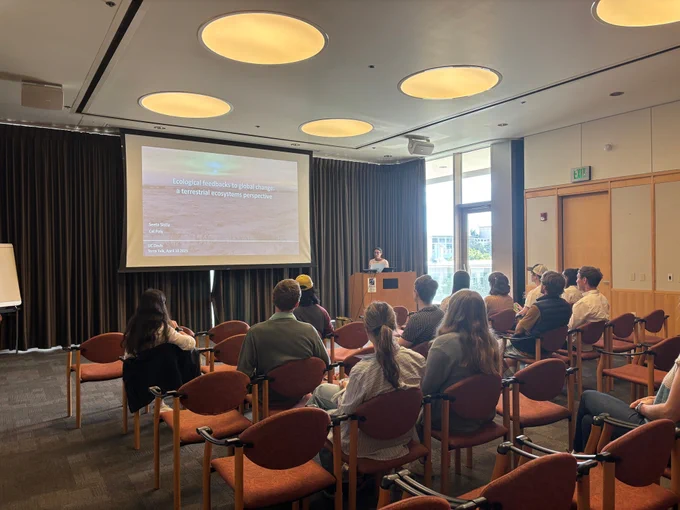 An audience faces a speaker at a podium who is presenting a presentation titled Ecological feedbacks to global change: a terrestrial ecosystems perspective.
