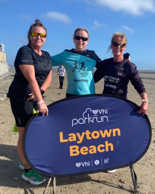 three parkrunners standing with their event sign at Laytown Beach parkrun