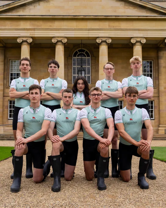 The Cambridge University Lightweight Men's Crew in white and Cambridge Blue and black wellies in front of one of Darwin College's buildings. They are arranged in two rows - five in the back with the cox, standing, and four kneeling on one knee in the front.