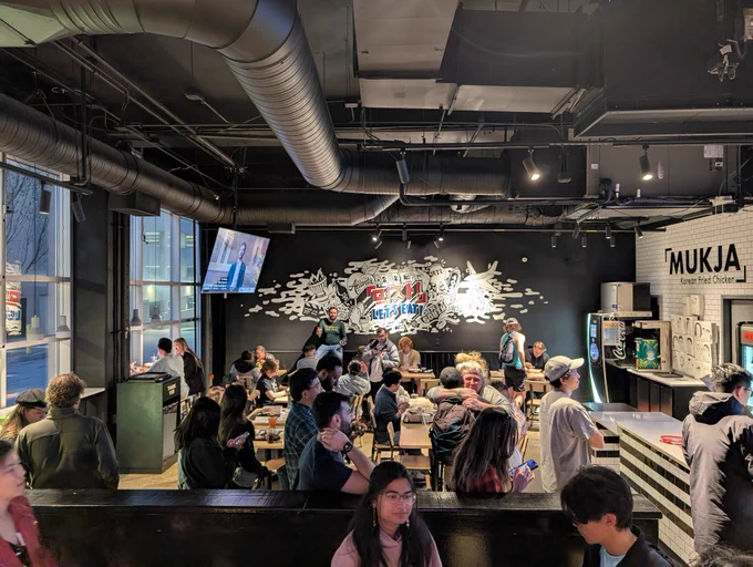Groups of people sitting at tables in a restaurant. Several people are standing in line.
