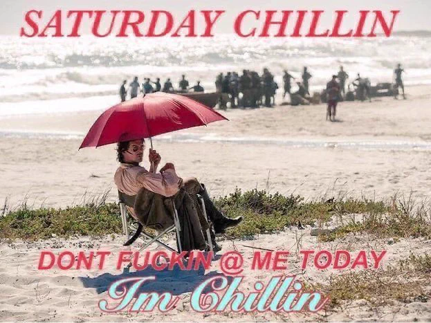 Toby Schmitz in costume as Jack Rackham from Black Sails is on a folding chair on a beach holding a small red umbrella on his head. Above him is red text in block capitals reads “Saturday chillin”. Below him there’s more text in the same capitals that reads “Don’t fuckin @ me today” and then blue cursive text that confirms: “I’m chillin”