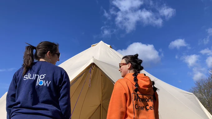 Two people smile at each other, facing the entrance to a bell tent, blue sky above them