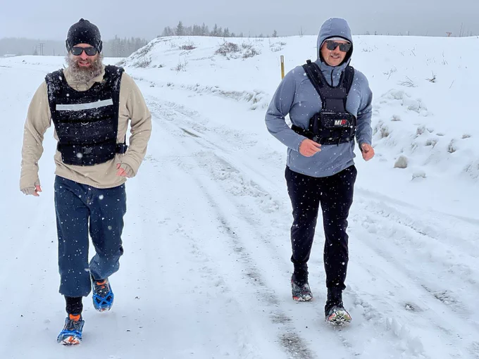 Two men wear black vests and athletic clothes as they walk along a snowy road while snow falls around them. 