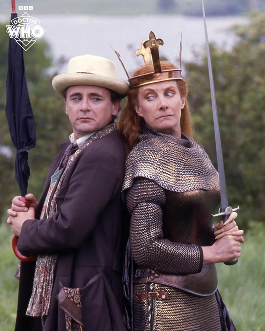 Jean Marsh as Morgaine in the Doctor Who episode 'Battlefield', brandishing a sword and standing back to back with Sylvester McCoy as the Seventh Doctor, who is brandishing an umbrella, they are both standing outside in a green field. 
