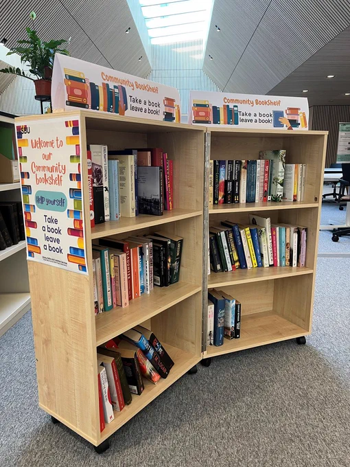 Photo taken showing a diagonal view of the Community Bookshelf. Double bookcase with three shelves on wheels with a selection of books on the shelves. 
