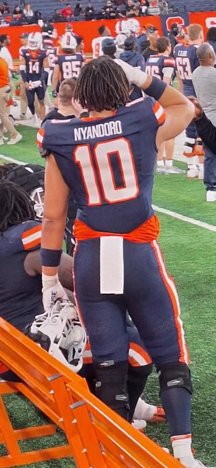 Nathan Nyandoro stands on the sideline with his right hand raised toward his head while standing next to teammates who are seated on a bench and behind another group of standing teammates on the sideline during the Syracuse Orange football team's spring game