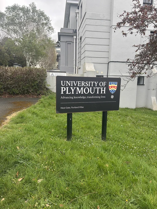 The University of Plymouth sign on a plot of grass with grey historical buildings in the background.
