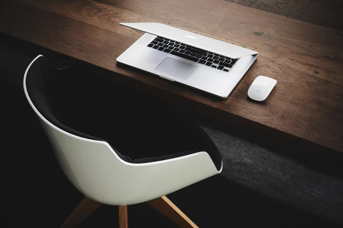 A modern workspace featuring an Apple MacBook and a white mouse on a wooden desk beside a stylish black and white chair.