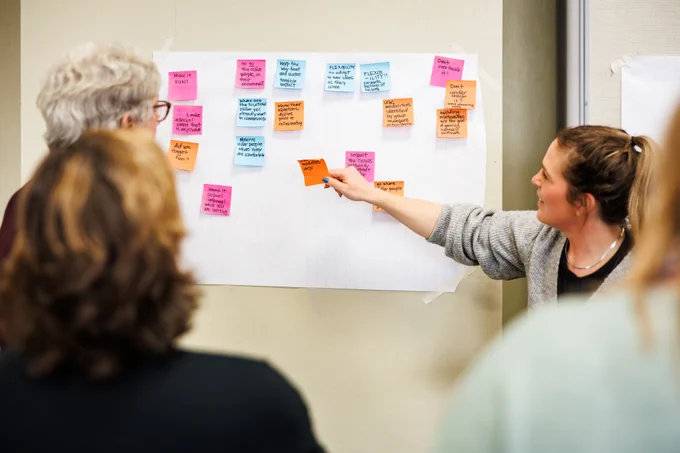 A student is pointing to a wall of post it notes with older adults, in conversation.