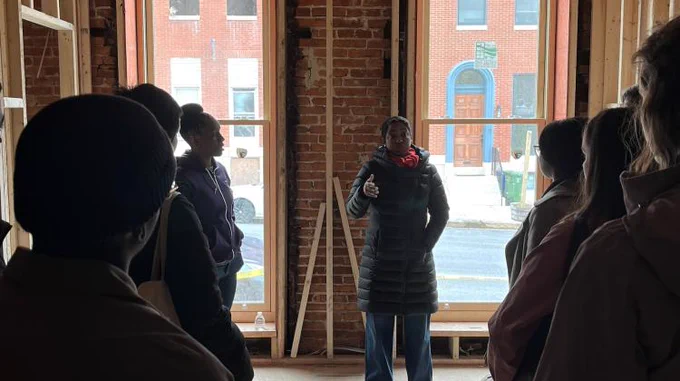 A group of students flank a pair of windows inside a townhouse. The building is stripped back to its studs.