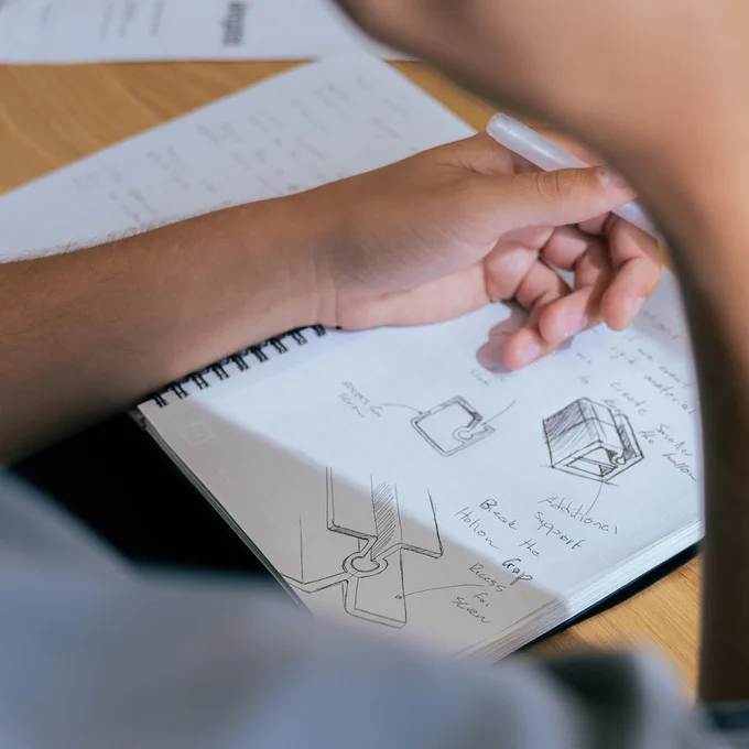 Person holding a pen and sketching product designs in a notebook on a wooden table.