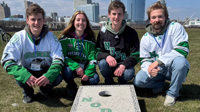 Three UND civil engineering students in hockey jerseys pose with a concrete cornhole board they constructed.