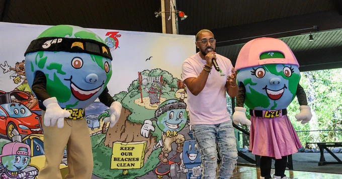 A 3rd Rock Hip Hop performer holds a microphone, flanked by two Earth-themed mascots, one wearing a black headband and the other in a pink baseball cap.