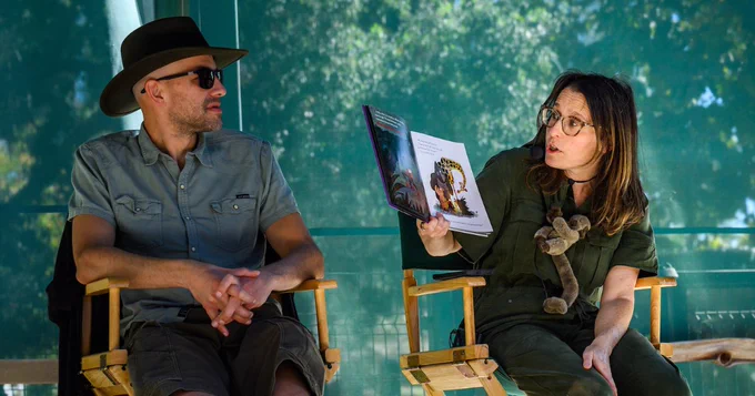Max and Suzanne Lang sitting on director's chairs, Max wearing a hat and looking at the Suzanne, who is holding and reading a Grumpy Monkey book.