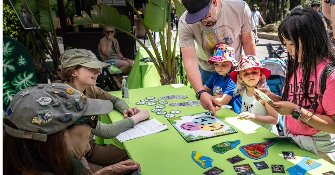 Adult and children engage in educational activities at a table covered with colorful animal illustrations and crafts, outdoors at an L.A. Zoo event.
