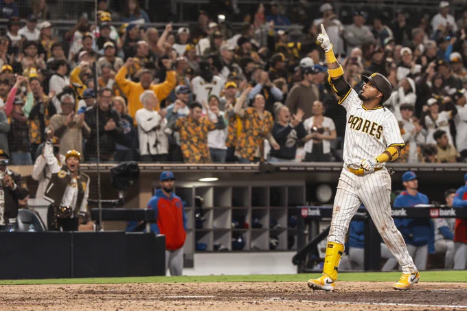 Luis Arraez about to step on home plate after hitting a home run and pointing up to the sky. Fans in the stands cheer him on in the background.