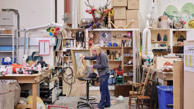Two individuals working in a cluttered art studio filled with various tools and materials. One is painting a figure, and the other is focused on paperwork at a desk.