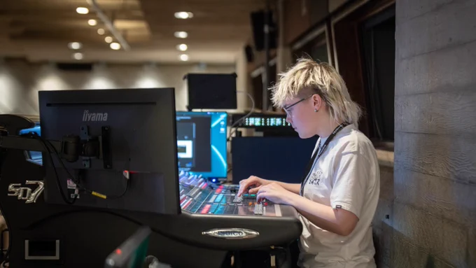 A person adjusts equipment in a room filled with monitors and technical gear.