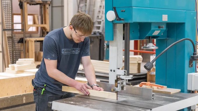 A worker is operating a large blue saw machine in a workshop, carefully guiding a piece of wood through it.