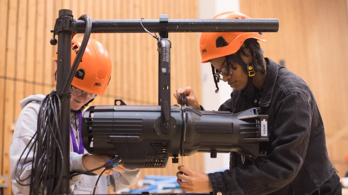 Two technicians in orange helmets are adjusting a large light fixture in a room with wooden paneling.