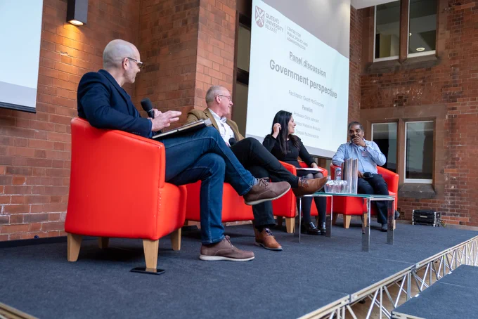 Image shows a panel discussion on stage. Geoff McGimpsey, the MC, is pictured along with Professor Chris Johnson, Professor Madalina Busuioc, and Dr Deepak Padmanabhan. 