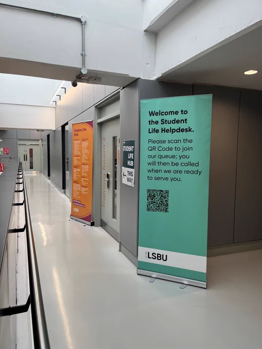 Photo of the outside of Training Room 2. Banner that advertises the support the Student Life Hub provides. Sign pointing to the door that reads 'Student Life Hub this way'. Banner with the QR code to join the queue.