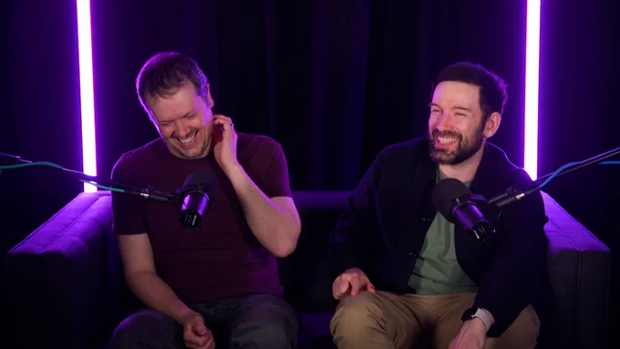 Mike and Andy laugh, sat together on a sofa with purple lighting behind them.
