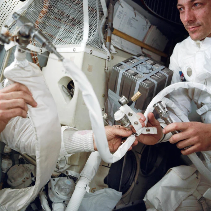 An interior view of the cramped Apollo 13 Lunar Module (LM) during the trouble-plagued journey back to Earth. This photograph shows some of the temporary hose connections and apparatus which were necessary when the three Apollo astronauts moved from the Command Module to use the Lunar Module as a “lifeboat.” Astronaut John L. Swigert Jr., command module pilot, is on the right. An unidentified astronaut on the left holds in his right hand the feed water bag from the Portable Life Support System (PLSS). Credit: NASA