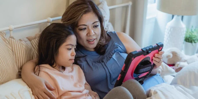 A mother and daughter share a moment, watching a tablet together in a cozy bedroom setting.