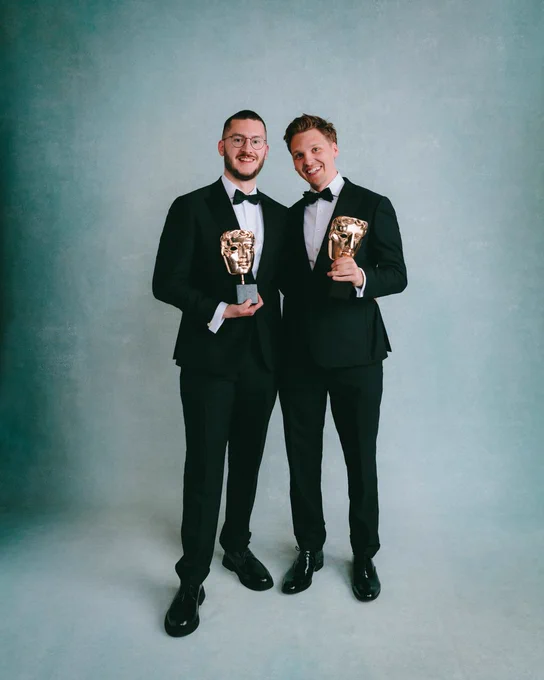 Will Todd and James Carbutt pose together with their BAFTA awards inside the BAFTA Games Awards portrait studio against a teal and white backdrop. They are both wearing black suits with bow ties, and stand shoulder to shoulder smiling for the camera and holding their awards in one hand. 