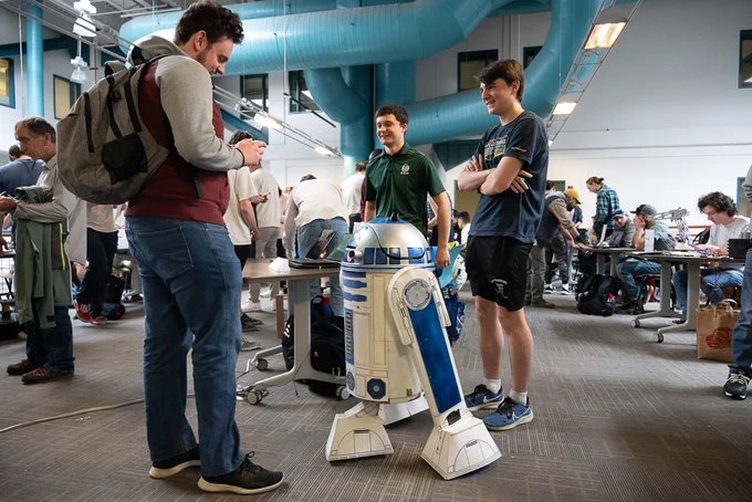 Three students interact with a life-sized R2-D2 replica at C S U’s E-Days event. One student takes a photo while the other two stand smiling near the droid in a busy indoor workspace filled with tables and attendees.

