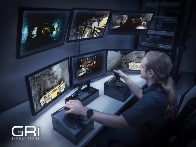 A man sits at a control station with two joysticks and six computer screens.