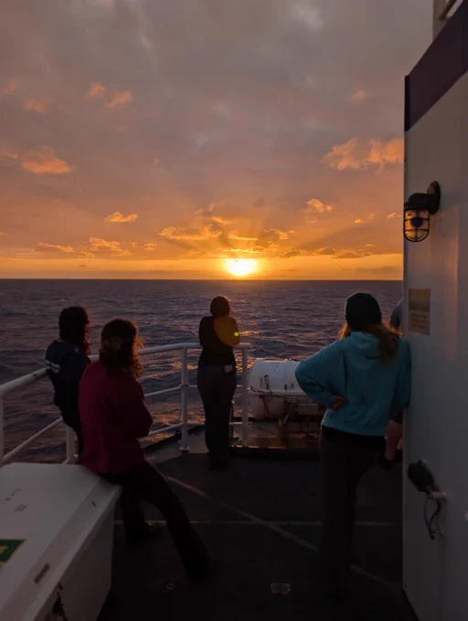 Four silhouetted people watch the sun set from the back deck of RV Sikuliaq.