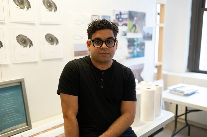 Photo by Rebecca Ou.
 
Description:
 
A headshot of a Puerto Rican man in an artist studio. He has short hair, a black shirt, and black rectangular glasses.
