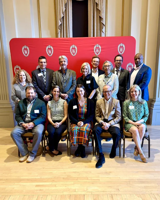 Group photo of Chancellor Mnookin and Provost Isbell with eleven Distinguished Teaching Award Recipients in front of a red Wisconsin crest backdrop.